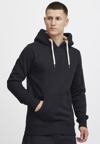Sudadera negra con capucha y bolsillo tipo canguro, cordones color crema y detalles en tono marrón alrededor de la capucha. Textura suave, puños y dobladillo acanalados.