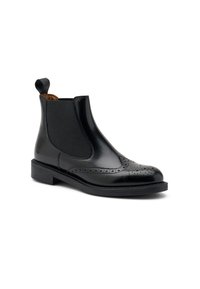 Botín Chelsea de cuero negro con detalles de brogue, paneles laterales elásticos, asa en la parte posterior y un tacón bajo sobre un fondo blanco.