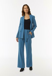 Turkusowy garnitur z dopasowaną marynarką i szerokimi spodniami. Gładki materiał oraz dwa guziki na marynarce. Czarna bluzka i buty na obcasie.