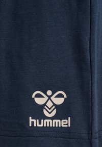 Logo Hummel w białym kolorze na granatowej tkaninie, przedstawiające stylizowaną pszczołę z napisem „hummel” pod spodem na materiale odzieżowym.