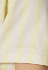 Vaaleanbeige kangas, jossa ohuet keltaiset raidat ja "LES DEUX" kirjailtuna valkoisella langalla helman läheisyydessä. Pehmeä, teksturoitu materiaali.