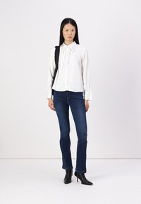 Blouse blanche à manches longues avec un col en nœud, associée à un jean fitted bleu foncé et des bottes à talons pointus noires. Design simple et épuré.