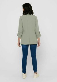 Frau mit schulterlangem, dunklem Haar, die ein hellgrünes Blazer, blaue Jeans und beigen Sneakers trägt und mit dem Rücken zu einem schlichten Hintergrund steht.