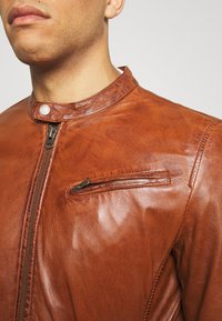 Giacca di pelle marrone con colletto alla mandarino, zip frontale e tasca sul petto con zip orizzontale. Texture liscia con una leggera lucentezza.