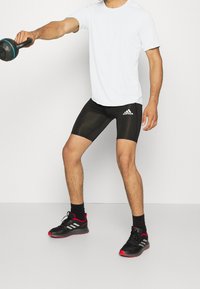 Camisa blanca de manga corta, pantalones cortos de compresión negros con logo blanco y zapatillas deportivas negras con acentos rojos; sosteniendo una pesa rusa.