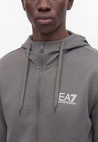 Gros plan d’une personne portant un sweat à capuche zippé gris Emporio Armani EA7 avec cordons et logo blanc sur la poitrine.