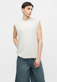 Jeune homme portant un débardeur blanc et un pantalon en denim bleu ample, debout devant un fond clair uni.