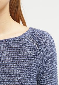 Gros plan sur une personne portant un pull lavande à maille lâche avec un motif texturé et de longs cheveux châtain roux visibles.