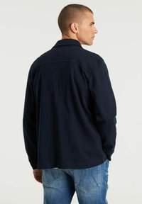 Marineblaue Langarmbluse mit Kragen, strukturiertem Stoff, einer Passe an der Rückseite und minimalem Markenlogo nahe dem Ausschnitt.