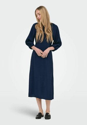 Blondína stojaca so zloženými rukami, oblečená v námornícky modrých midi šatách s nafúknutými rukávmi a čiernych nazúvacích topánkach.