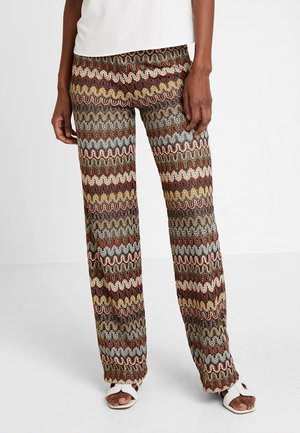 Persoon met wijde broek met meerkleurig zigzag haakwerkpatroon, gecombineerd met witte open-toe sandalen en een wit topje zichtbaar bij de taille.