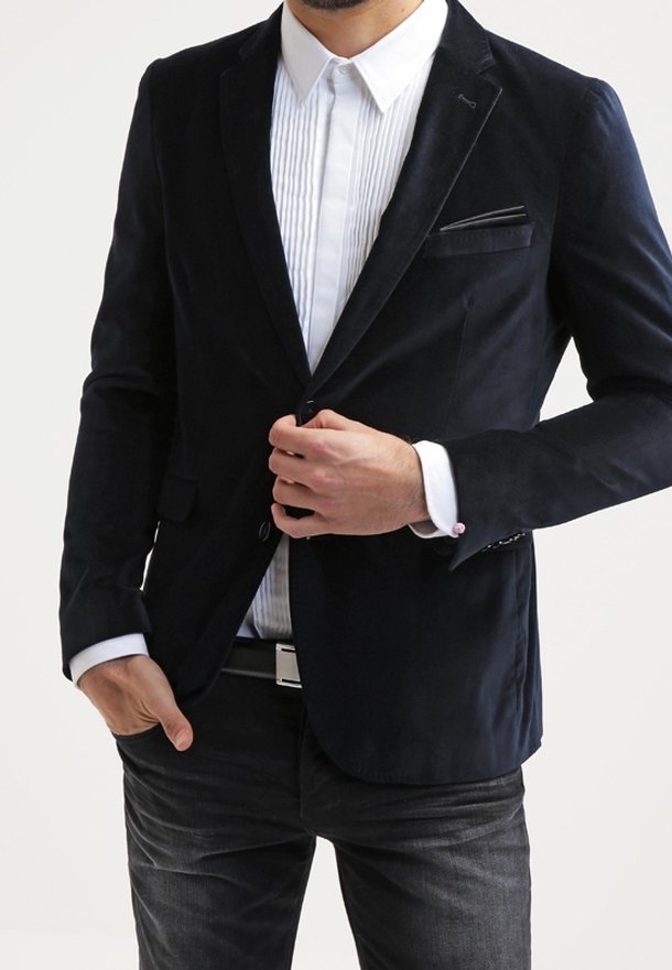 Blazer di velluto a coste blu navy con vestibilità slim, tasca singola e una texture sottile. Indossato sopra una camicia bianca plissettata e jeans scuri.