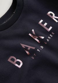 Marineblaues Sweatshirt mit glänzenden, reflektierenden Buchstaben. Der Text lautet "BAKER by TID BAKER" und ist auffällig über den Stoff verteilt.