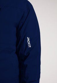 Marineblå jakke med et glat, struktureret stof. "JACK1T"-logoet er tydeligt præsenteret i hvid på venstre ærme.