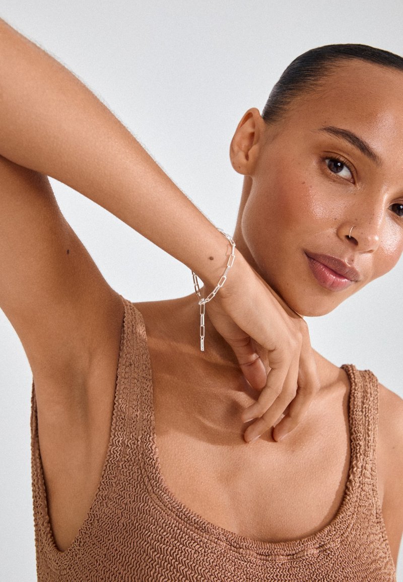 Hõbedahela käevõru, millel on piklikud lingid ja ripats, kantud pruuni tekstuuriga toppen, esitledes minimalistlikku disaini.