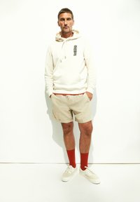 Felpa crema con grafica nera, pantaloni corti beige, calzini rossi e sneakers bianche. Look casual con vestibilità rilassata e design semplice.
