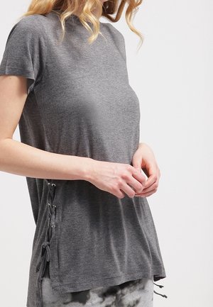 Frau trägt ein locker sitzendes graues Kurzarmoberteil mit seitlichen Schnürdetails, steht vor einem schlichten hellen Hintergrund.