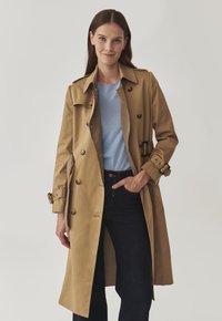 Tan trench coat with double-breasted button closure, epaulets, and side pockets, layered over a blue shirt and dark jeans.