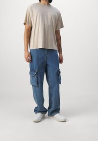 Beige bomulds T-shirt med løs pasform, kombineret med blå denim cargo bukser med to sidelommer, afsluttet med hvide sneakers.