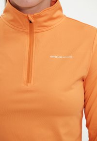 Oranje kwart-rits pullover gemaakt van rekbare stof. Voorzien van een opstaande kraag en een wit merkembleem op de borst. Gladde textuur.