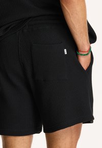 Fekete, waffle-rácsos anyagú shorts, textúrált felülettel, oldalsó zsebbel és derékrésszel, hátul egy kis márkajelzéssel.