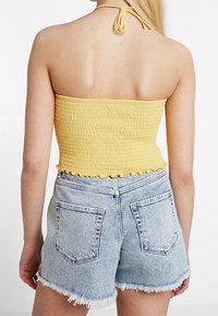 Žena oblečená v žlutém halter topu s nařasením a světle modrých roztřepených džínech, viděná zezadu na bílém pozadí.