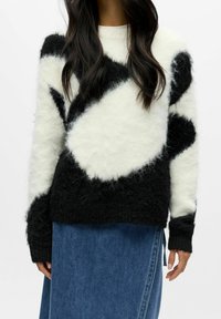 Kvinne iført en svart og hvit genser med ullaktig mønster og blå denimskjørt, stående mot en ensfarget bakgrunn.