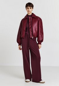 Giubbotto bomber in pelle bordeaux con maniche a sbuffo, abbinato a pantaloni a gamba larga coordinati, caratterizzati da coulisse e dettagli nelle cuciture laterali.