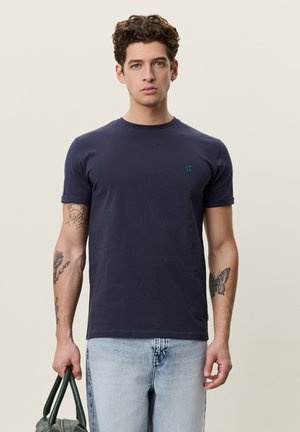 Młody mężczyzna z kręconymi włosami, ubrany w ciemnogranatowy T-shirt i jasne dżinsy, trzymający zielone uchwyty torby, z tatuażami na obu przedramionach.