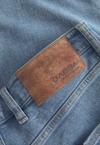 Jeansy dżinsowe w jasnoniebieskim kolorze z fakturowaną brązową skórzaną łatą z napisem "GABBA". Charakteryzują się starannym przeszyciem i klasycznym designem z pięcioma kieszeniami.