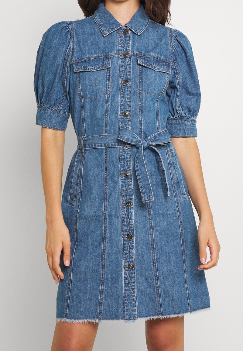 Mukena denim dengan lengan puff, kerah, depan kancing, dua saku dada, tali pinggang, dan hem yang robek. Warna biru muda dan tekstur halus.
