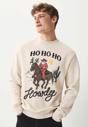 Ung man som bär en beige sweatshirt med jultomten i rött ridande på en ren dekorerad med ornament, kaktusar, öken och texten "HO HO HO Hej."