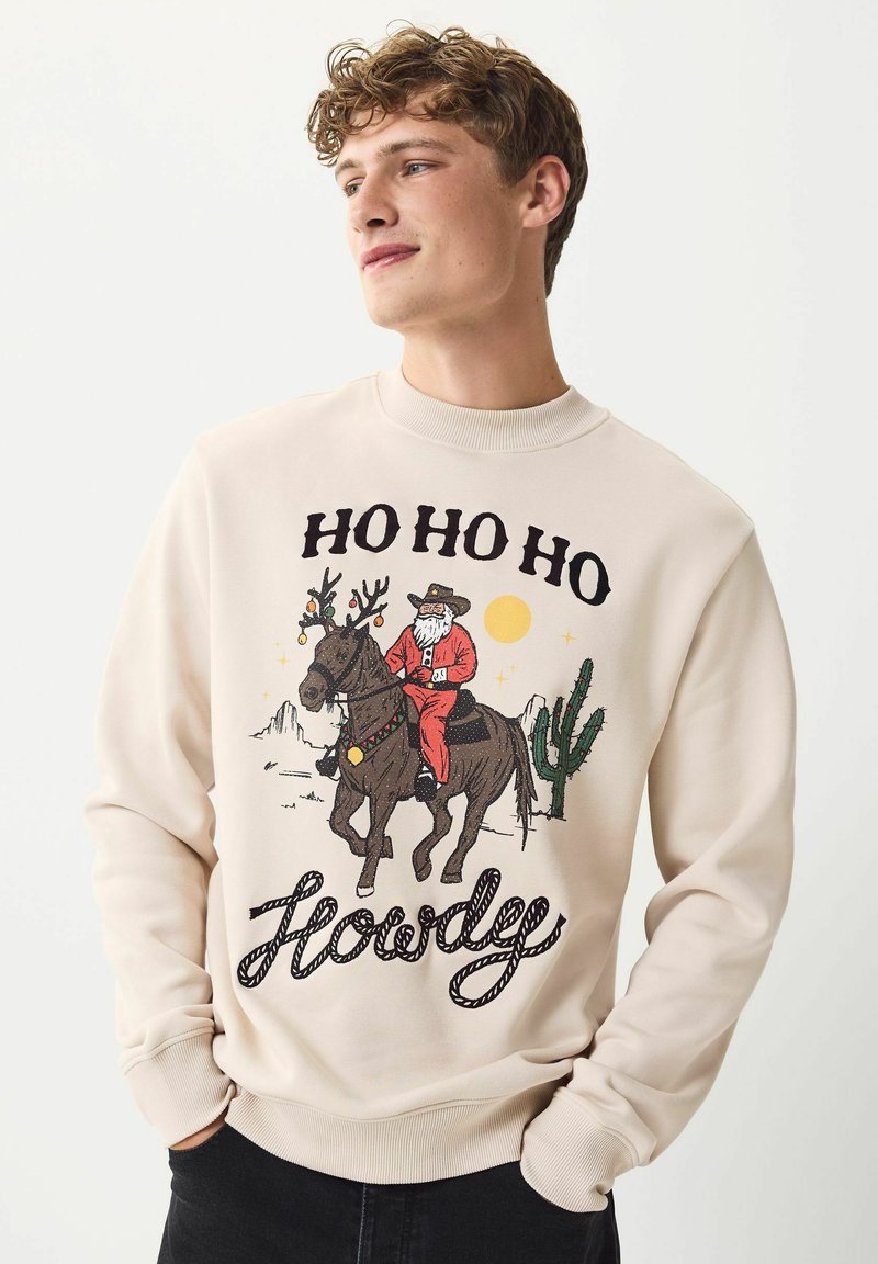 Ung man som bär en beige sweatshirt med jultomten i rött ridande på en ren dekorerad med ornament, kaktusar, öken och texten "HO HO HO Hej."