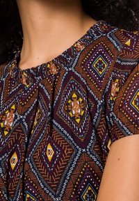 Cou du et épaules d'une personne portant un haut à manches courtes avec un motif géométrique en losanges multicolores et un décolleté froncé.