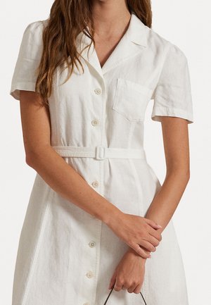 Femme portant une robe blanche à manches courtes boutonnée avec une ceinture, tenant un petit sac, bras croisés devant.