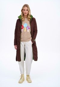 Brązowy pikowany długi płaszcz z zielonymi akcentami, beżowy sweter z kolorowymi wzorami grzybów, białe spodnie i beżowe botki.