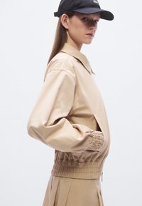 Μπεζ bomber μπουφάν με γιακά, ελαστική μέση και σούρες στα μανίκια. Φορεμένο με μαύρο καπέλο που έχει λογότυπο στο μπροστινό μέρος.