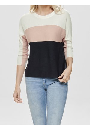 Femme portant un pull color block avec des sections crème, rose et noire, associé à un jean bleu clair, sur un fond uni.