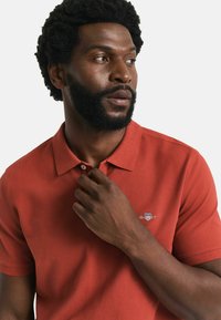 Rotes Poloshirt aus strukturiertem Stoff mit einem Kragen mit zwei Knöpfen, kurzen Ärmeln und einem kleinen gestickten Logo auf der Brust.