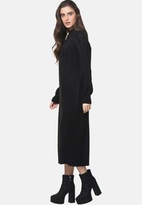 Robe en tricot noire avec des manches longues, un col montant et une texture côtelée. Portée avec des bottines à talons hauts noires.