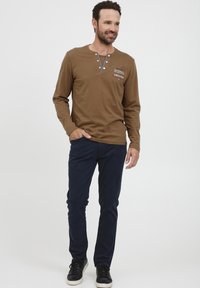 Brun langermet skjorte med henley-hals, blå detaljer og logo, parret med mørk marineblå bukser og sorte joggesko.