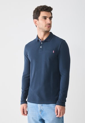 Polo de manga larga en color navy, hecho de tela suave, con una tapeta de dos botones y un bolsillo con un acento de rayas rojas y blancas.