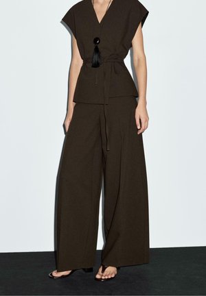 Femme portant un top cache-cœur sans manches marron foncé et un pantalon à jambes larges avec un collier pendentif noir et des sandales à bouts ouverts sur un fond uni.