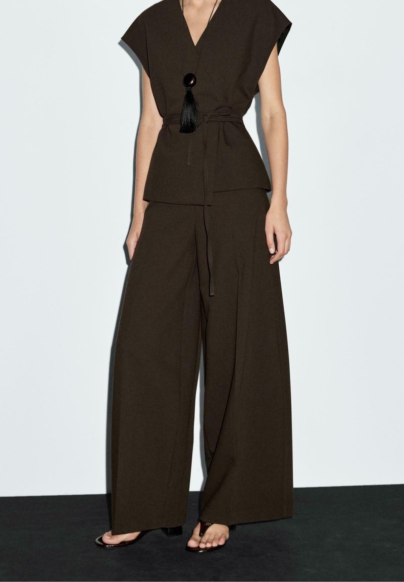 Femme portant un top cache-cœur sans manches marron foncé et un pantalon à jambes larges avec un collier pendentif noir et des sandales à bouts ouverts sur un fond uni.