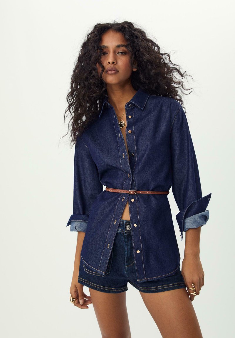 Dunkelblaue Jeanshemd mit langen Ärmeln, Knopfverschluss und kontrastierenden Nähten. Kombiniert mit Jeansshorts und einem braunen Gürtel.