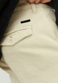 Pantaloni in corduroy beige con sottili righe verticali, dotati di una tasca laterale con bottone e un'etichetta nera vicino alla vita.