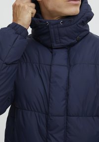 Marineblå puffjakke med høy krage, trykknapper og justerbar snor. Kviltet tekstur, med sidelommer.