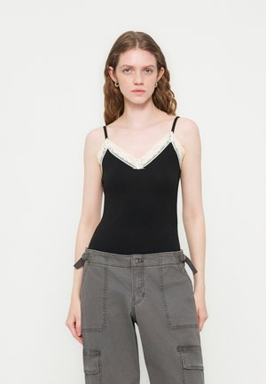 Jeune femme aux cheveux ondulés portant un caraco noir avec bordure en dentelle blanche et un pantalon cargo gris sur fond blanc uni.