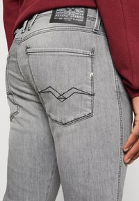 Jeans de denim gris con un corte ajustado, que presentan costuras decorativas en los bolsillos traseros y una etiqueta de cuero en la cintura con detalles de la marca.