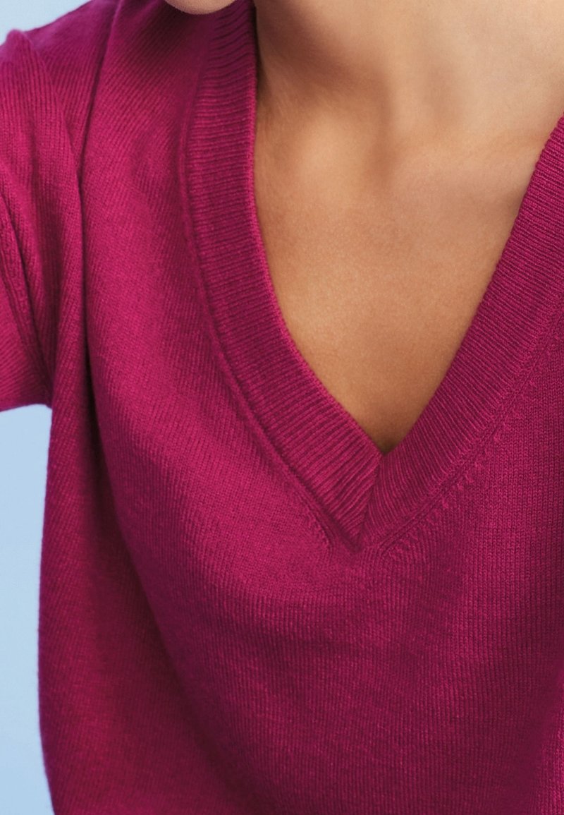 Pull col en V couleur fuchsia avec une texture en maille fine et douce. Comprend des poignets et un ourlet côtelés. Vue rapprochée mettant en valeur le détail de l'encolure et des manches.